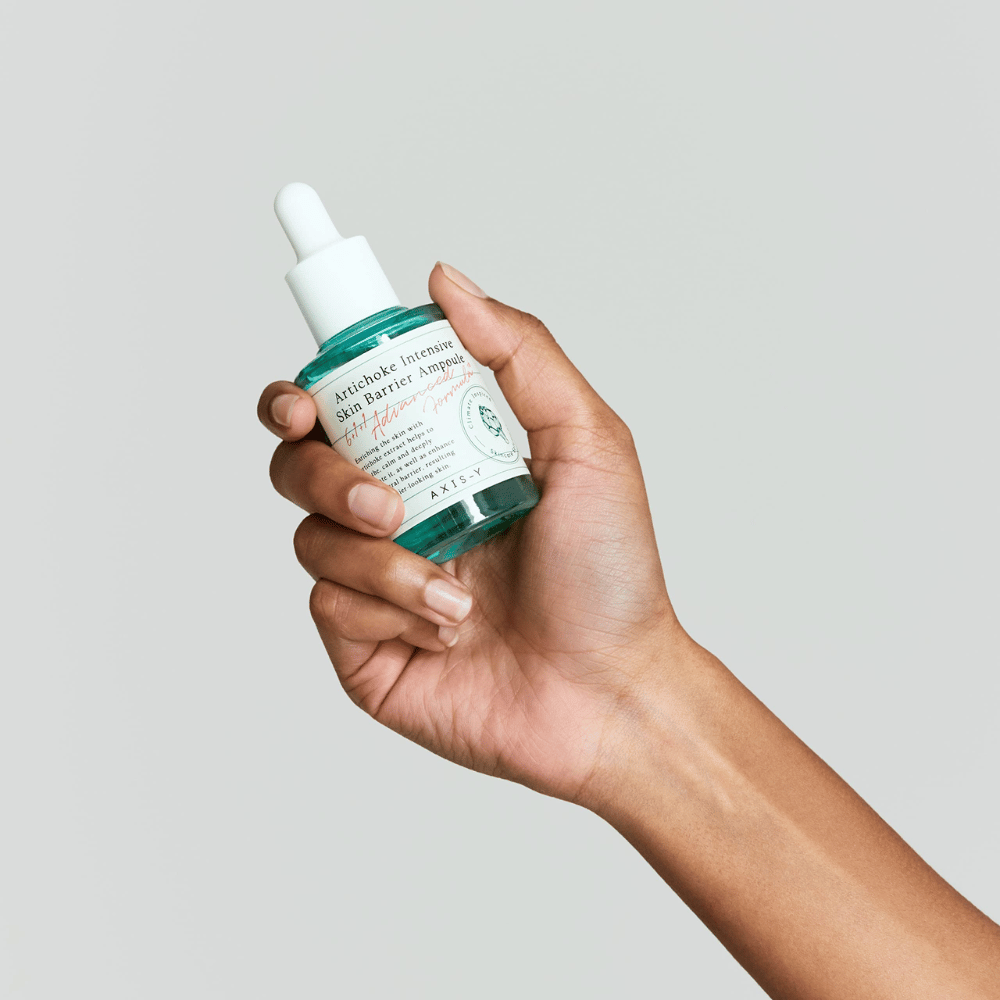 En hand håller en flaska hudvårdsserum med etiketten "Artichoke Intensive Skin Barrier Ampoule.