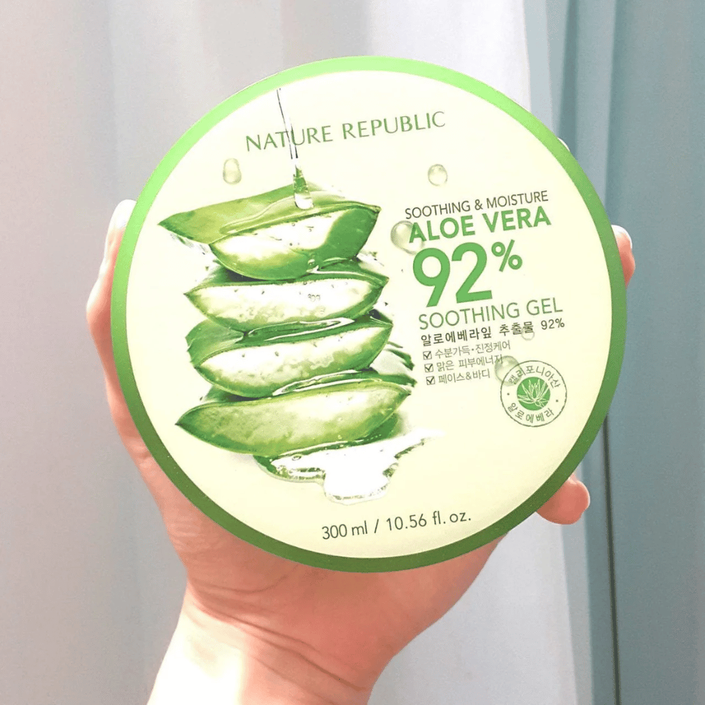 En hand håller upp en rund burk med Nature Republic Aloe Vera 92% Soothing Gel. Burken är grön med en bild av aloe vera och text som beskriver dess lugnande och fuktgivande egenskaper. Innehållet är 300 ml.