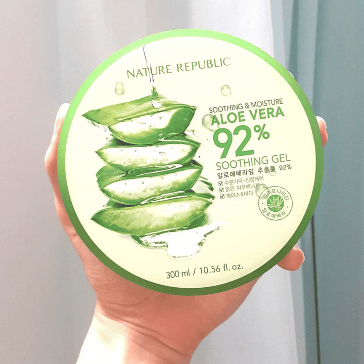 En hand håller upp en rund burk med Nature Republic Aloe Vera 92% Soothing Gel. Burken är grön med en bild av aloe vera och text som beskriver dess lugnande och fuktgivande egenskaper. Innehållet är 300 ml.